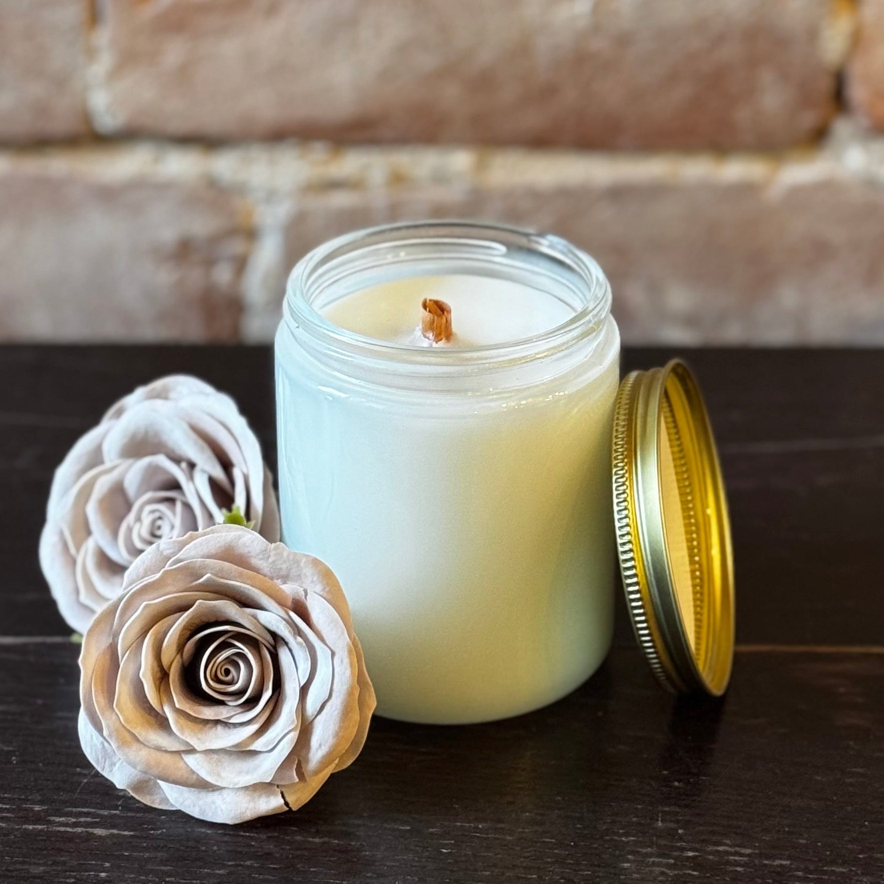 A custom candle with a wooden wick in an 8oz container, surrounded by decorative flowers set against a brick wall backdrop.