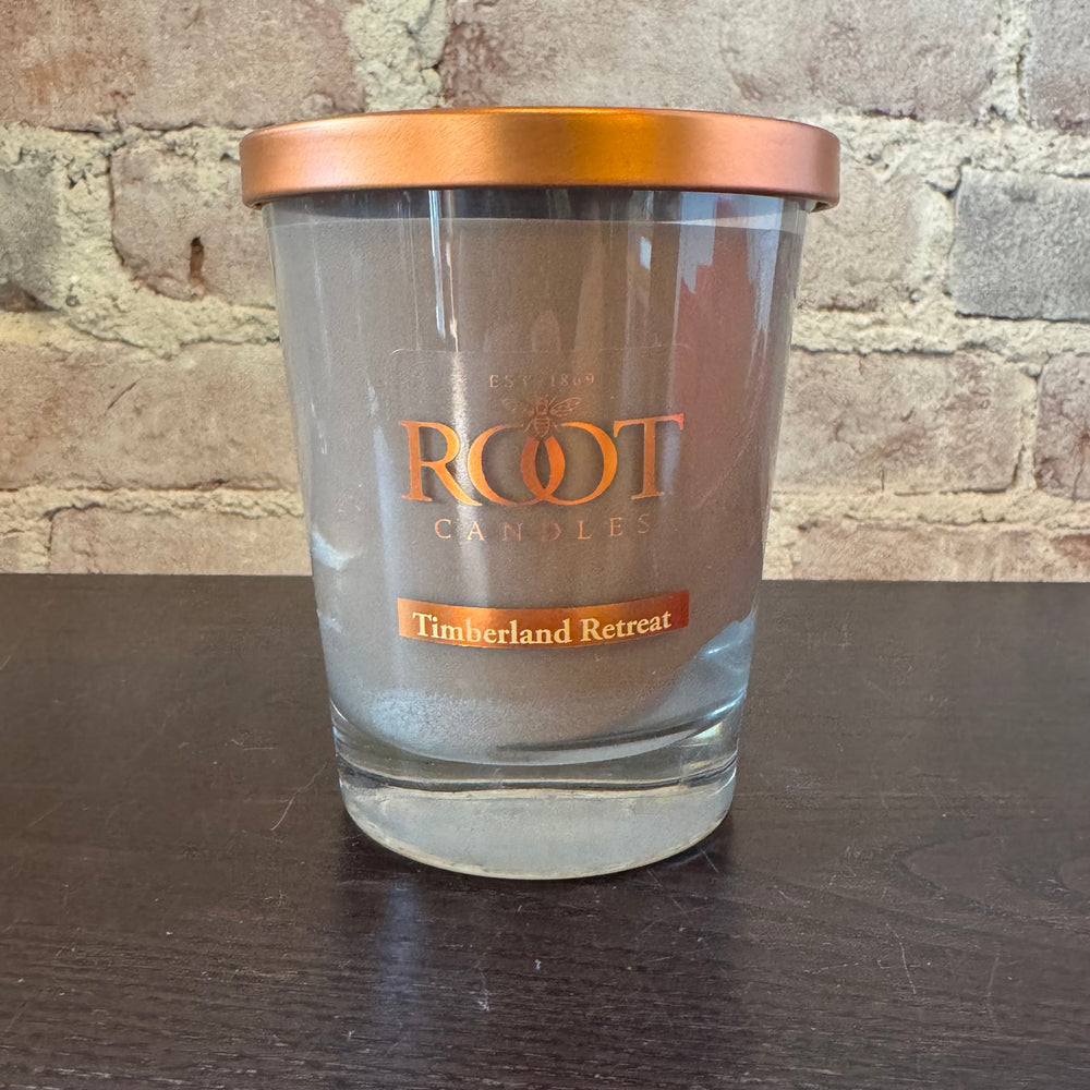 Glass candle with copper lid labeled 'Root Candles Timberland Retreat' on a wooden surface.