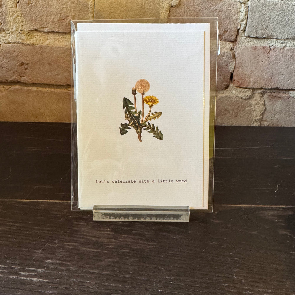 Greeting card with dandelion flowers and text 'Let's celebrate with a little weed' on a stone surface.