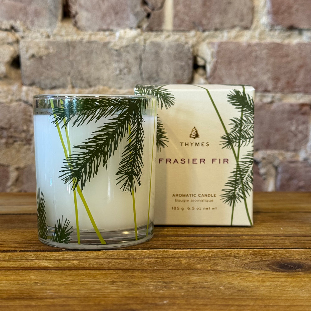 Thymes Frasier Fir candle and packaging on a wooden surface with a brick wall background