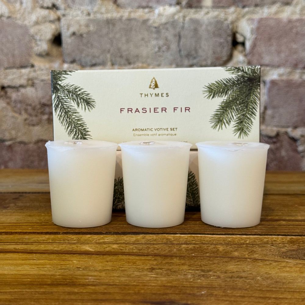 Three Thymes Frasier Fir votive candles on a wooden surface with packaging in the background.