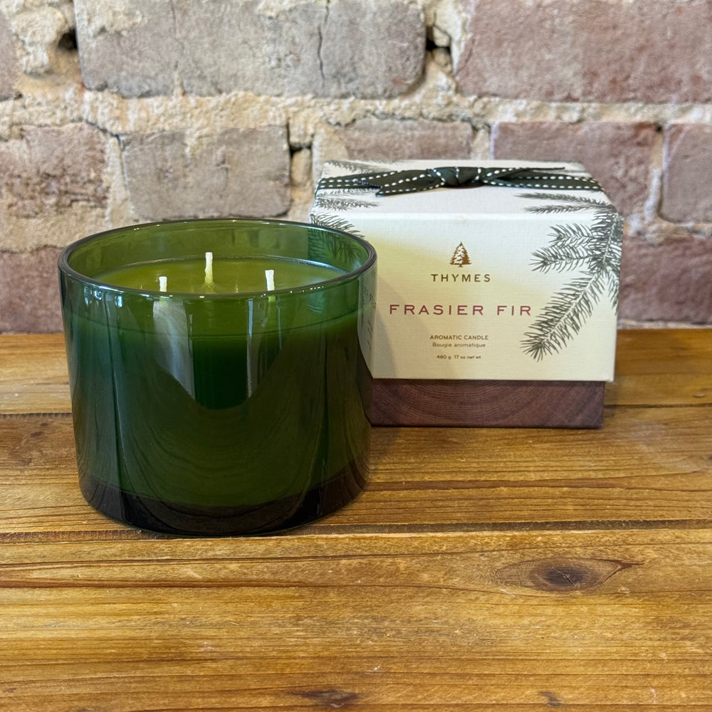 Green candle in a glass jar with a Thymes Frasier Fir candle box on a wooden surface.