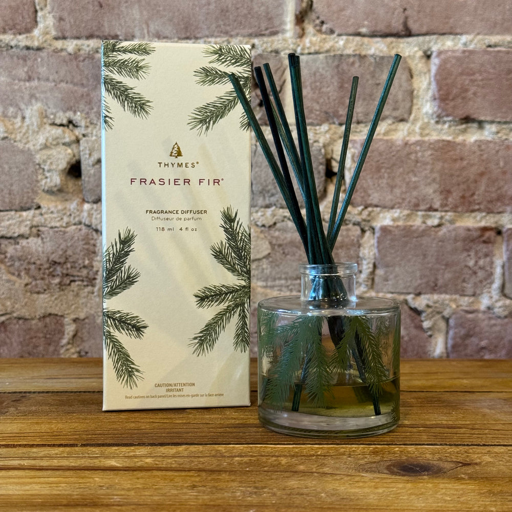 Thymes Frasier Fir fragrance diffuser with packaging on a wooden surface against a brick wall.