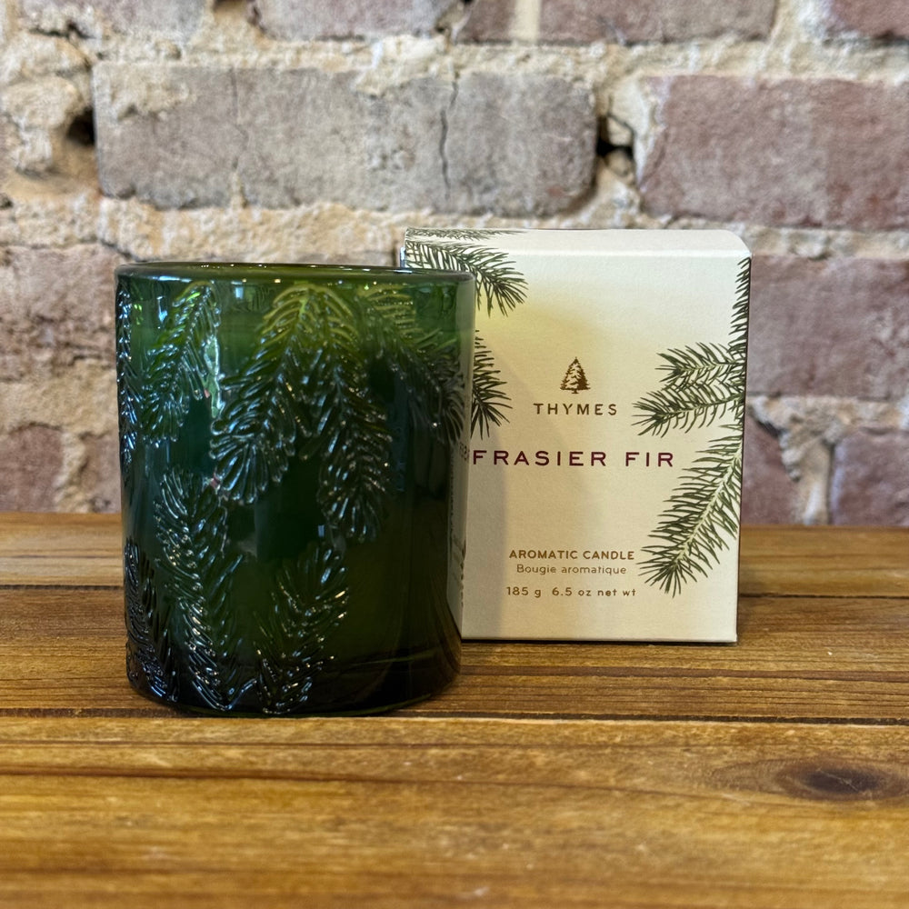 Green textured candle with Thymes Frasier Fir packaging against a brick wall.