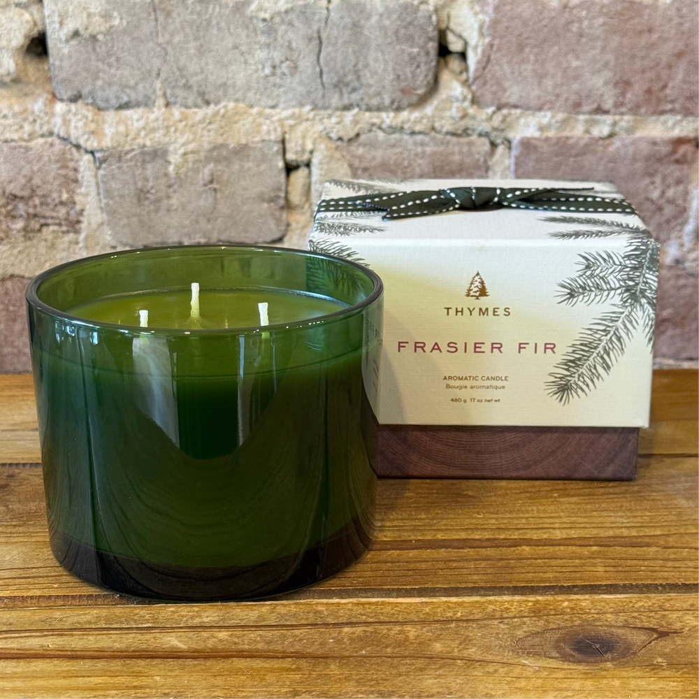 Candle in a green glass jar with Thymes Frasier Fir packaging on a wooden surface.