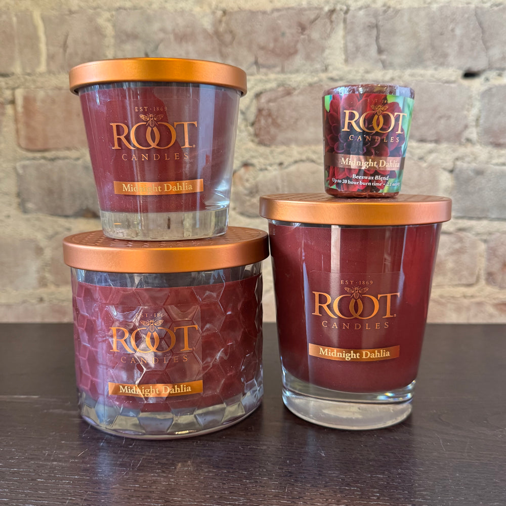 Three Root Candles labeled 'Midnight Dahlia' on a wooden surface with a brick wall background.