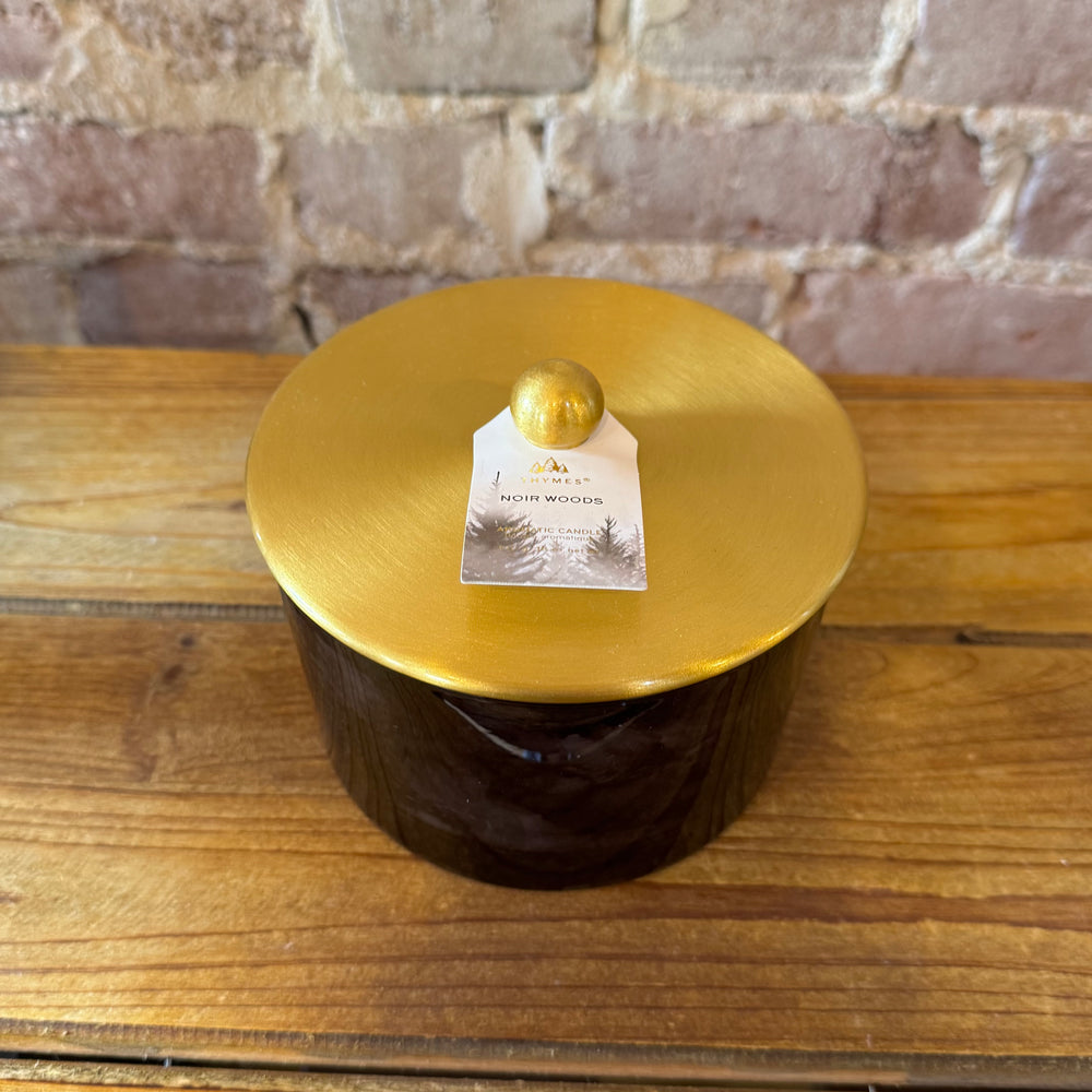 Top view of black candle jar with gold lid and wooden base against a brick wall.