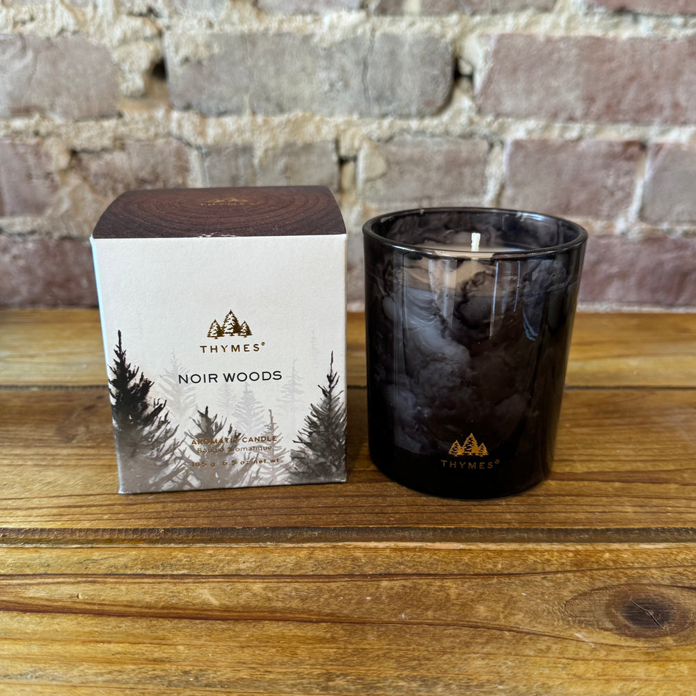 Thymes Noir Woods candle and packaging on a wooden surface with a brick wall background