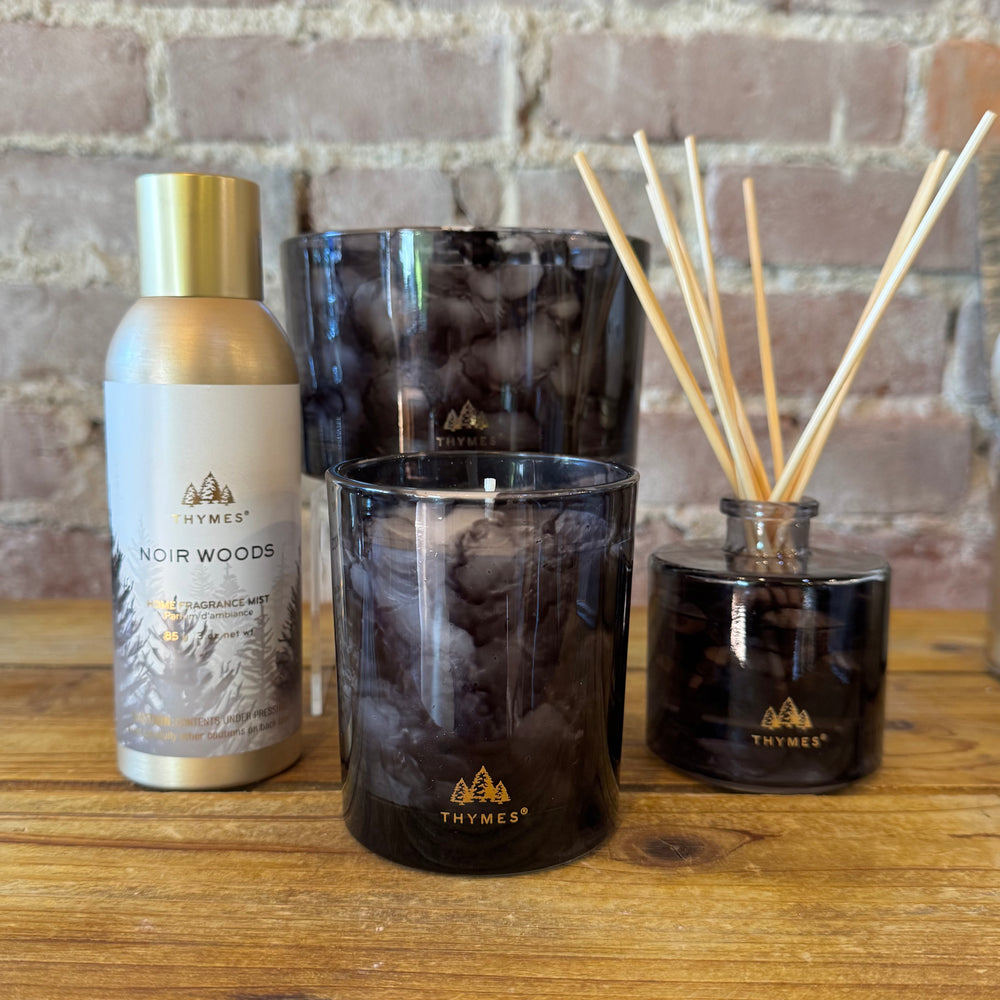 Thymes Noir Woods fragrance products including a bottle, diffuser, and candle on a wooden surface with a brick wall background.