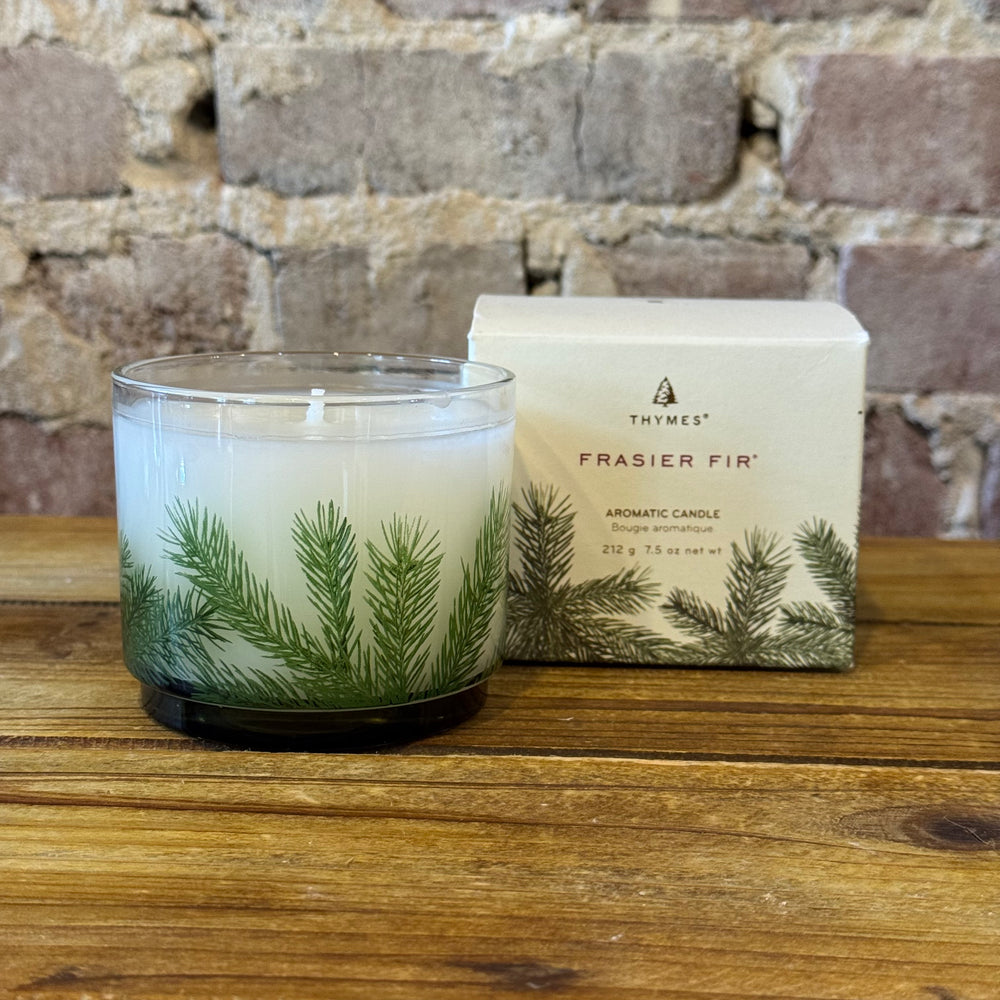 Thymes Frasier Fir candle and packaging on a wooden surface with a stone wall background